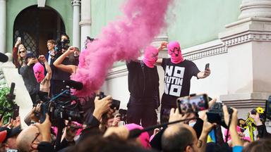 Schwarz gekleidete Menschen mit pinkfarbenen Gesichtsmasken protestieren vor einem grünen Gebäude, während eine Menge aus Reportern und Zuschauern, filmt und fotografiert.