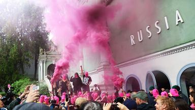 Schwarz gekleidete Menschen mit pinkfarbenen Gesichtsmasken protestieren vor einem grünen Gebäude, während eine Menge aus Reportern und Zuschauern, filmt und fotografiert.