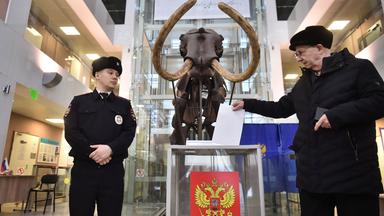 Ein Mann steckt ein Blatt Papier in eine große Wahlurne aus durchsichtigem Plexiglas mit dem russischen Wappen. Daneben steht ein Mann in Uniform und schaut zu, im Hintergrund ist das Skelett eines Mammuts in einem naturhistorischen Museum zu sehen.
