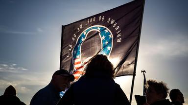Vor einer von der untergehenden Sonne angeleuchteten Flagge mit einerm großen Q in den Farben der amerikanischen Flagge, sind die Silhouetten mehrerer Personen dunkel zu sehen.