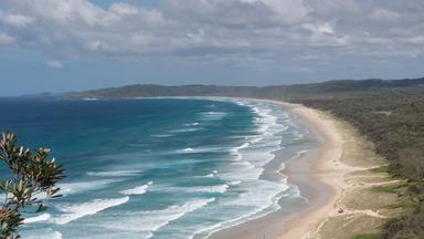 Panoramaaufnahme eines weitläufigen Sandstrandes der in ein dichtes Waldgebiet übergeht. Am blauem Himmel hängen einige Wolken.