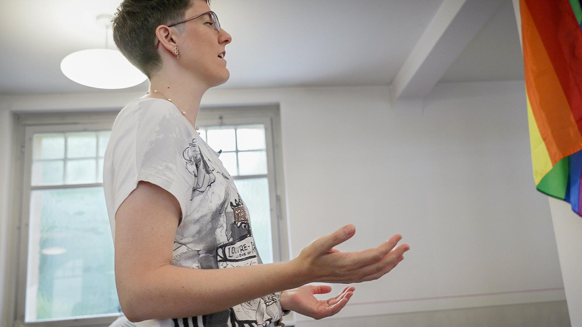 Eine Person mit Kurzhaarfrisur steht in einem Raum mit Regenbogenflagge und spricht gestikulierend, vermutlich im Rahmen eines Gesprächs oder Vortrags.