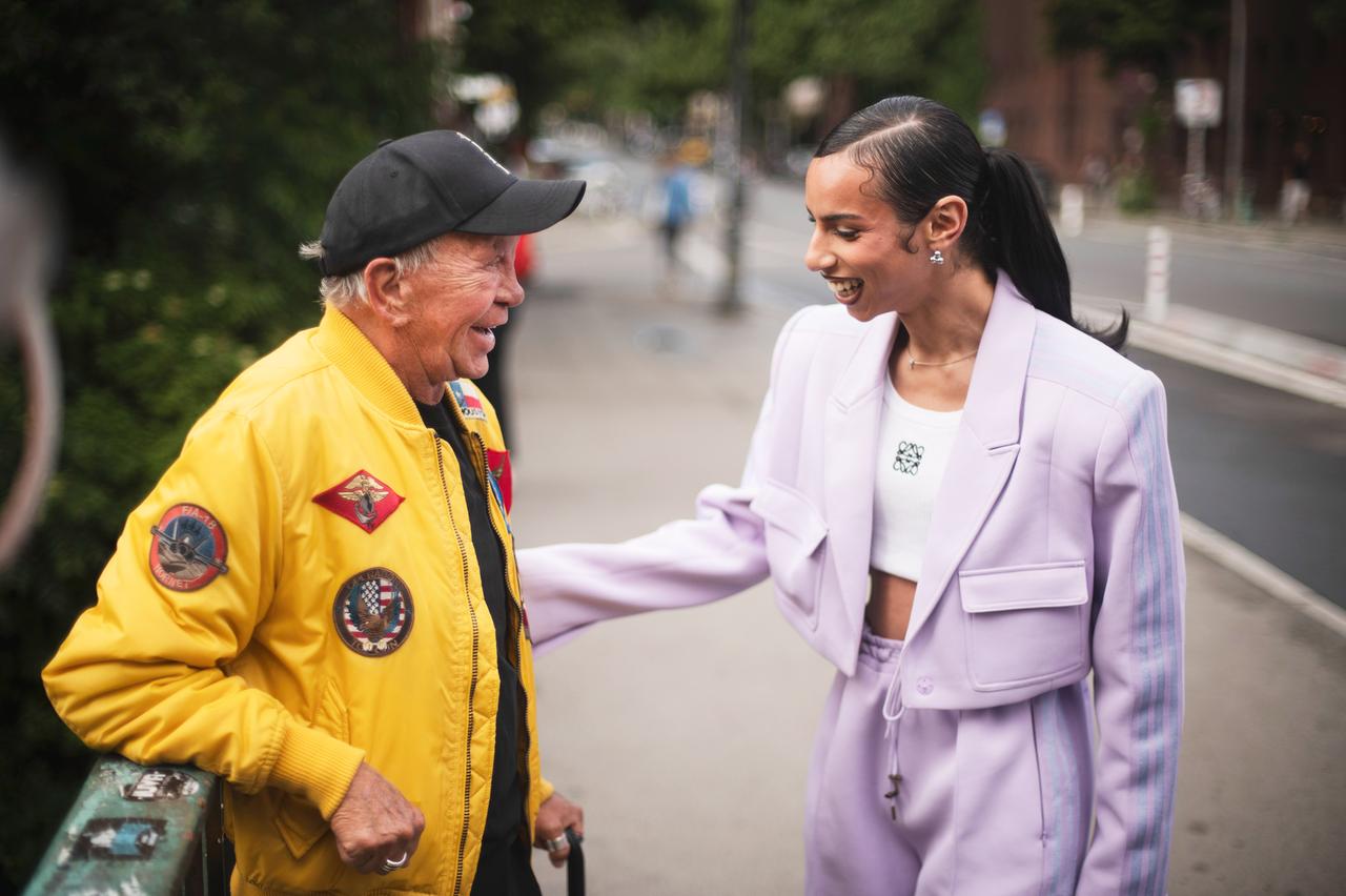 Ein älterer Mann in gelber Jacke und schwarzer Kappe steht lachend einer jungen Person in einem hellvioletten Hosenanzug gegenüber. Beide unterhalten sich auf einer Straße in Berlin.