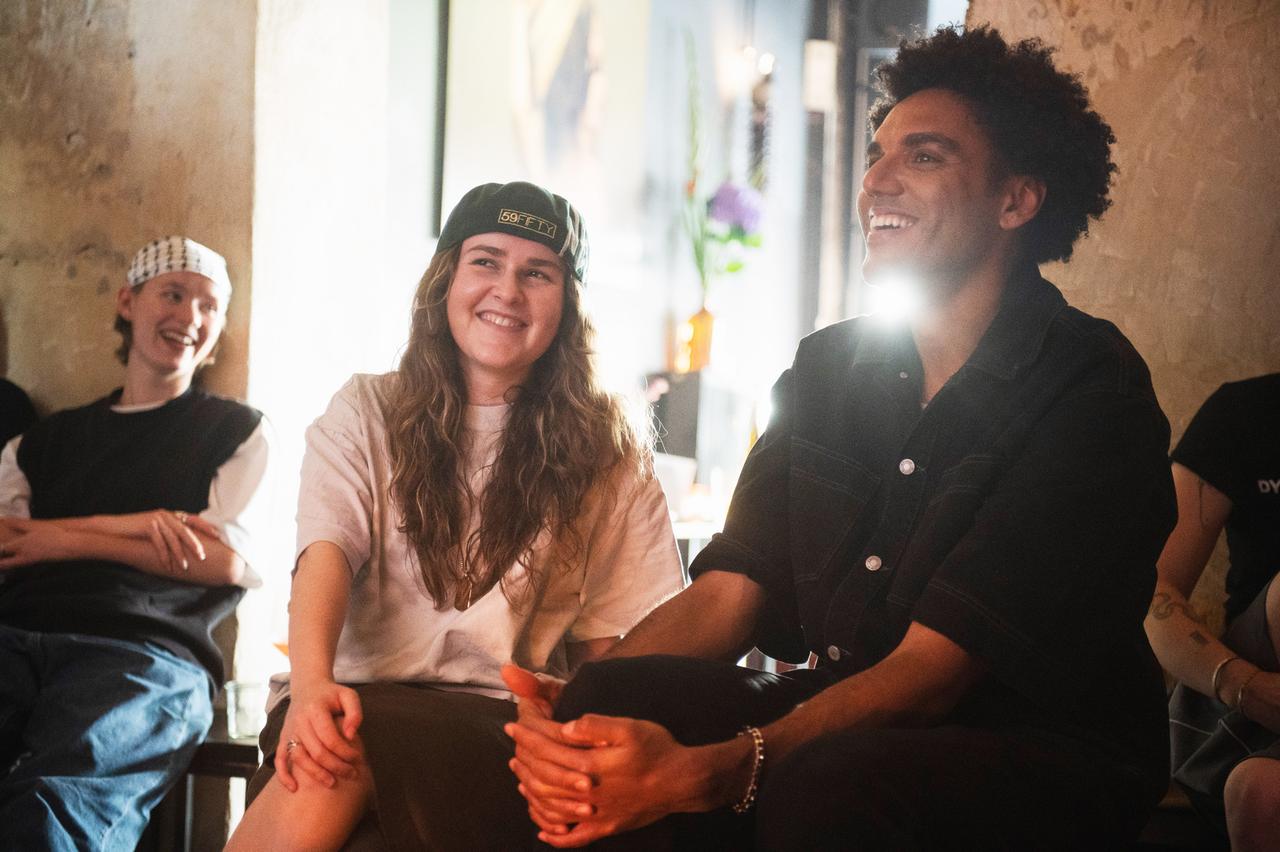 Eine junge Frau und ein junger Mann sitzen nebeneinander in einem Raum mit warmem Licht und lachen. Die Frau trägt ein helles T-Shirt und eine Basecap, der Mann ein dunkles Hemd. Im Hintergrund sitzen weitere Menschen und unterhalten sich.