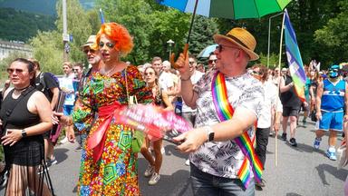 Das Bild zeigt eine Szene von einer Pride Parade in Innsbruck, Österreich. Im Vordergrund sind mehrere Teilnehmer zu sehen, die fröhlich marschieren. Eine Person trägt ein auffälliges, buntes Kleid mit geometrischem Muster sowie eine orangefarbene Perücke und hat eine bewundernde Gestik. Neben ihr ist ein weiterer Teilnehmer, der ein Hemd mit tropischem Muster trägt und ein Regenbogen-Sternenbanner um die Schulter geworfen hat.   Der Mann hält einen grünen Regenschirm und hat eine fröhliche Miene. Im Hintergrund sind eine große Menschenmenge zu sehen, die ebenfalls feierlich und ausgelassen agiert. Viele tragen bunte Accessoires, wie Regenbogenflaggen oder geschmückte Kleidung. Die Straße ist gesäumt von Bäumen, die eine grüne Umgebung bieten. Die Atmosphäre wirkt festlich und voller Freude, was die Botschaft von Toleranz und Diversität unterstreicht. Der gesamte Anlass vermittelt ein Gefühl von Gemeinschaft und Unterstützung für die LGBTIQ+-Bewegung.