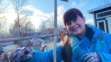 Sabrina Nitsche steht mit warmer Winterkleidung und Handschuhen im Garten und hält lächelnd einen gefrorenen Beerenzweig in der Hand.