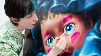 Frau mit kurzen Haaren und blauer Strähne malt mit Pinsel an einem großen puppenartigen Gesicht auf Leinwand.