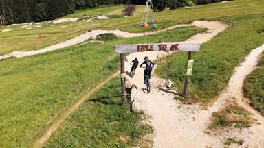 Das Bild zeigt einen Trailpark in der Oststeiermark, Österreich. Im Vordergrund steht ein Holzschild mit der Aufschrift "FREE TO BE". Der Hintergrund ist geprägt von saftig grünen Wiesen, die mit kleinen Hügeln und Erdwegen durchzogen sind. Zwei Personen auf Mountainbikes fahren auf einem geschotterten Pfad, der von der beschilderten Stelle abgeht. Im Hintergrund sind weitere Radfahrer zu sehen, die den Trail nutzen. Links im Bild ist eine Rückwärtsbahn zu erkennen, die zum Transport von Radfahrern dient. Die Umgebung ist von Bäumen umgeben, die eine natürliche Kulisse bieten. Das Bild vermittelt eine aktive, naturnahe Atmosphäre des Radfahrens in einer malerischen Landschaft.