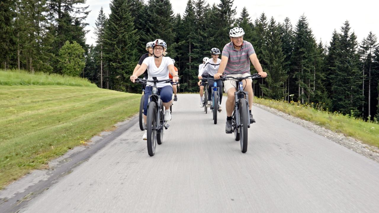Das Bild zeigt eine Gruppe von sechs Personen, die auf Fahrrädern eine asphaltierte Straße entlangfahren. Die Radfahrer tragen Helme und unterschiedlich farbige Sportbekleidung. Die Landschaft um sie herum ist geprägt von grünen Wiesen und Wäldern. Auf der linken Seite des Bildes sind hohe, grüne Bäume zu sehen. Der Himmel ist teilweise bewölkt, und die Straße ist flach und gut befahrbar. Die Szene vermittelt einen Eindruck von Outdoor-Aktivität und Naturverbundenheit in der Oststeiermark, Österreich.