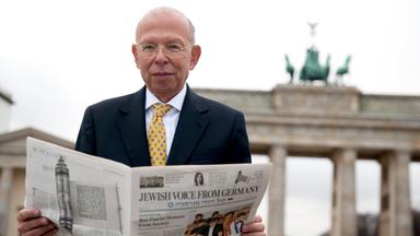 Rafael Seligmann hält Zeitung in der Hand - im Hintergrund das Brandenburger Tor