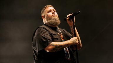 Das Bild zeigt einen männlichen Musiker, der auf einer Bühne auftritt. Er hat einen rasierten Kopf mit lockigen Haaren an den Seiten und einen dichten, grauen Bart. Sein Oberteil ist schwarz, und er trägt einen kurzen, ärmellosen Überwurf. Auf seinem linken Arm sind auffällige Tattoos sichtbar. In seiner rechten Hand hält er ein Mikrofon und singt mit ernstem Gesichtsausdruck. Der Hintergrund des Bildes ist dunkel und leicht verschwommen, was die Bühne und die Lichtverhältnisse während des Auftritts betont. Das Bild vermittelt eine energiegeladene Atmosphäre eines Live-Konzerts.