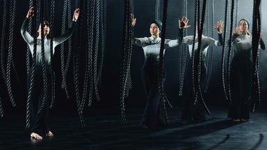 Bühnenszene: Frauen stehen mit erhobenen Händen zwischen herabhängenden Seilen.