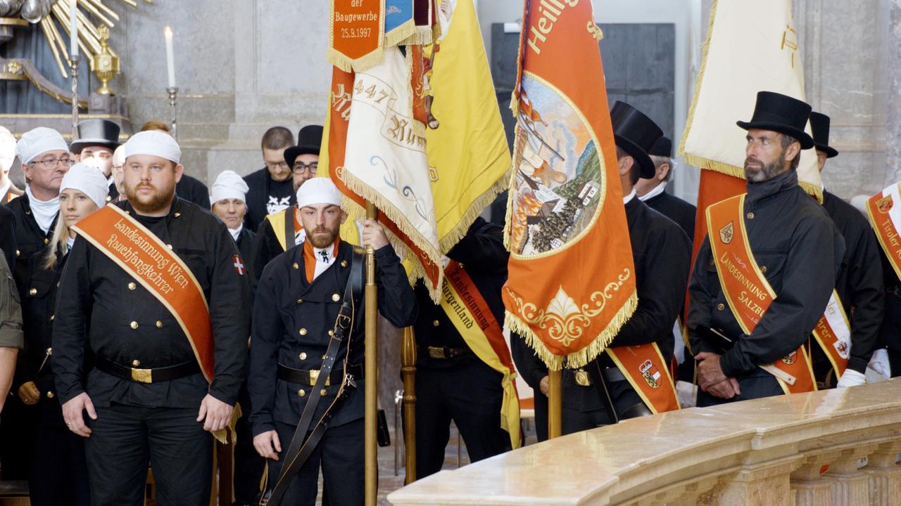 Das Bild zeigt eine Gruppe von Rauchfangkehrern, die in einer festlichen Umgebung versammelt sind. Sie tragen traditionelle schwarze Arbeitskleidung, mit goldenen Knöpfen und weißen Kopfbedeckungen. Einige von ihnen haben Schärpen mit Aufschriften, die auf die „Landesinnung Wien“ hinweisen. In der Mitte des Bildes steht ein Raucherkehrer, der eine Fahne hält, auf der verschiedene Wappen und Symbole abgebildet sind.   Im Hintergrund sind weitere Personen zu sehen, die ebenfalls festliche Kleidung tragen und aufmerksam in die gleiche Richtung schauen. Die Umgebung wirkt feierlich und traditionell, mit Dekorationen, die auf ein besonderes Ereignis hindeuten. Die Beleuchtung ist hell und hebt die Details der Kleidung und der Fahnen hervor.  Das Bild vermittelt ein Gefühl von Gemeinschaft und traditioneller Handwerkskunst, während die Rauchfangkehrer ihrer Verantwortung als Glücksbringer und Handwerker nachgehen.
