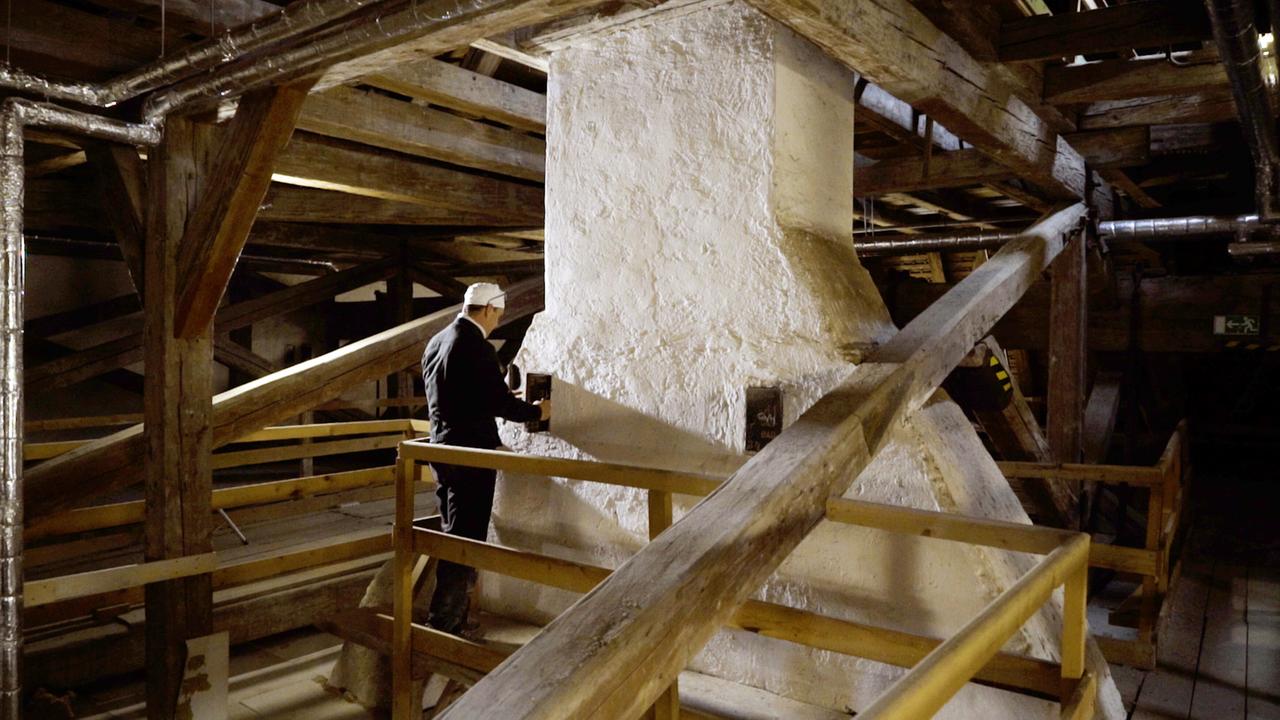 Das Bild zeigt einen Dachboden der Orangerie des Schlosses Schönbrunn in Wien. Im Raum sind zahlreiche Holzstrukturen sichtbar, darunter dicke Balken, die das Dach stützen. In der Mitte des Bildes steht ein Rauchfangkehrer, der schwarz gekleidet ist und eine Kappe trägt. Er steht an einer großen, weißen Wand, die sich als Kamin herausstellt. Der Rauchfangkehrer berührt offensichtlich die Oberfläche mit beiden Händen und scheint eine Prüfung oder Wartung durchzuführen. Um ihn herum sind Holzgeländer sichtbar, die den Arbeitsbereich abgrenzen. Über dem Kamin sind metallische Rohre sichtbar, die eine technische Installation andeuten. Das Licht im Raum ist gedämpft, was eine atmosphärische Stimmung erzeugt.