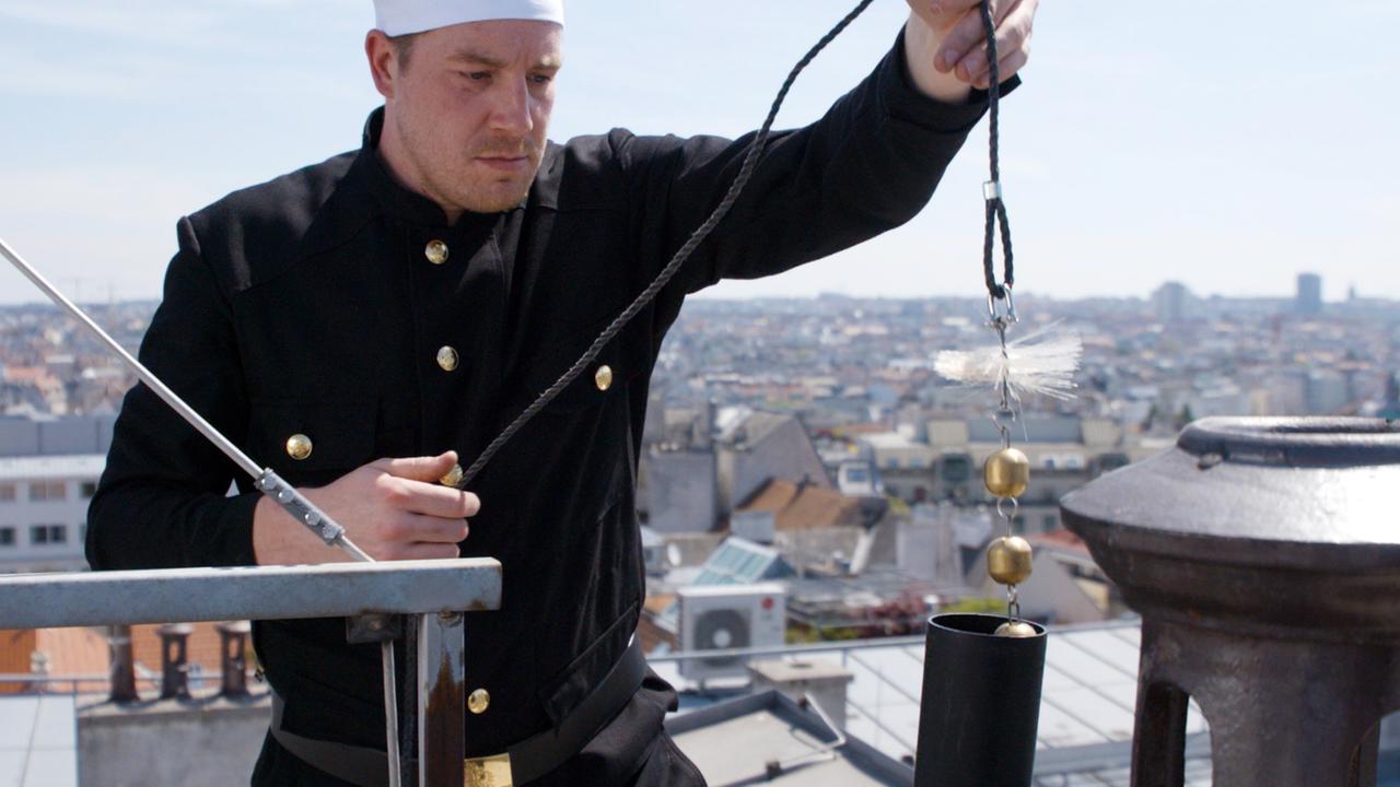 Das Bild zeigt einen Rauchfangkehrer, der auf einem Dach arbeitet. Er trägt eine schwarze Arbeitsjacke mit goldenen Knöpfen und eine weiße Kopfbedeckung. In seiner rechten Hand hält er ein Werkzeug, das aus einem langen Seil mit einer Bürste an einem Ende besteht. Das andere Ende des Seils ist mit einer goldenen Kugel verbunden.   Im Hintergrund erstreckt sich eine städtische Skyline, die aus verschiedenen Gebäuden und Dächern besteht. Die Szene ist bei Tageslicht aufgenommen, und der Himmel ist klar. Die Umgebung deutet auf eine große Stadt hin, wahrscheinlich Wien, da der Film über die Wiener Rauchfangkehrer handelt.   Insgesamt vermittelt das Bild den Eindruck von handwerklicher Tradition und moderner Arbeitsweise in einem urbanen Umfeld.
