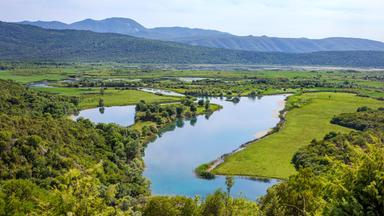 Eine Flusslandschaft mit grünen Auen und Hügeln im Hintergrund. Klare Gewässer schlängeln sich durch die saftig bewachsenen Ebenen.