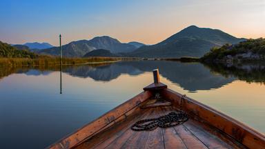 Eine Holzbootspitze auf einem stillen See, umgeben von Bergen im Hintergrund, in Montenegro bei Sonnenuntergang.