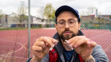 Ein Mann steht vor einem Sportplatz und zerreißt einen Joint.