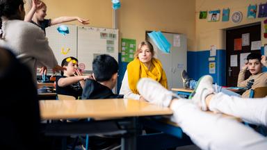 Lara sitzt inmitten einer Schulklasse. Schülerinnen und Schüler haben die Füße auf den Tischen und werfen Papierflieger durch den Klassenraum.
