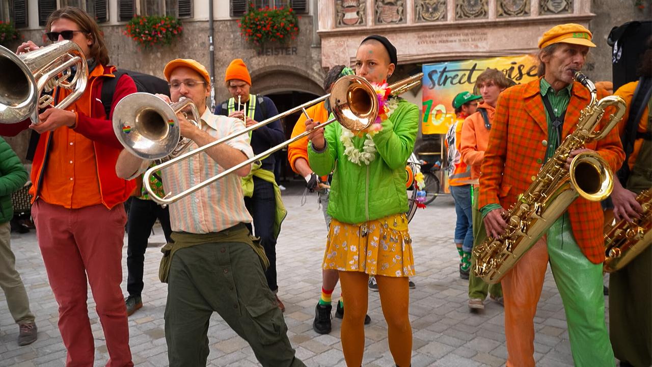 Das Bild zeigt eine lebendige Szene eines musikalischen Auftritts. Eine Gruppe von Personen steht auf einem gepflasterten Platz, umgeben von historischen Gebäuden. Die Musiker halten verschiedene Blasinstrumente, darunter Trompeten und Posaunen. Die Kleidung der Musiker ist auffällig bunt: Grüntöne, Orange, Gelb und rot dominieren.   Die zentrale Person, die Posaune spielt, trägt eine grüne Jacke und einen Kranz aus Blüten. Neben ihr steht ein weiterer Musiker in einer gestreiften Bluse und einer beigen Hose. Im Hintergrund sind weitere Mitglieder der Gruppe zu sehen, die ebenfalls verschiedene Instrumente spielen. Ein Banner im Hintergrund kündigt "street noise" an und unterstreicht den urbanen Charakter der Aufführung.   Die Szene vermittelt eine Atmosphäre von Freude und Kreativität, typisch für Straßenkunst und Musikdarbietungen in städtischen Umgebungen.