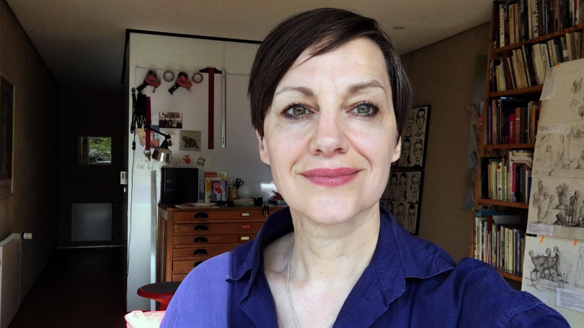 Porträtbild einer Frau, die in einem Zimmer mit Bücherregal im Hintergrund steht.