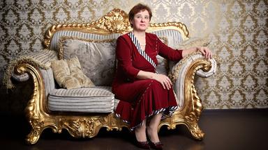 Eine Frau in einem roten Kleid sitzt steif und aufrecht auf einem vergoldeten Sofa vor einer mit goldener Brokattapete