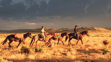Zwei Menschen reiten auf Pferden durch die Landschaft. Im Hintergrund ziehen dunkle Wolken auf.