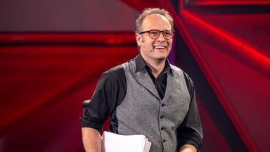 René Sydow ist in der Mitte des Bildes zu sehen. Er trägt einen Stapel Papier unter dem rechten Arm. Der Hintergrund ist rot beleuchtet.