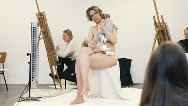 Sitzend posierende Frau mit Babypuppen auf dem Schoß und im Arm, umringt von Malenden an Staffeleien.