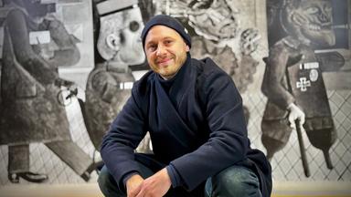 Ein Mann sitzt in Hockstellung vor einem Gemälde und lächelt in die Kamera. Er trägt eine dunkelblaue Mütze und Jacke und eine Jeans. Das Gemälde hinter ihm ist ein großformatiges schwarz-weiß Kunstwerk.