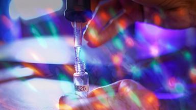 Nahaufnahme einer Pipette, die eine klare Flüssigkeit in ein kleines beschriftetes Fläschchen tropft; eine Hand hält das Gefäß, bunte Lichtreflexe überlagern die Laborszene.
