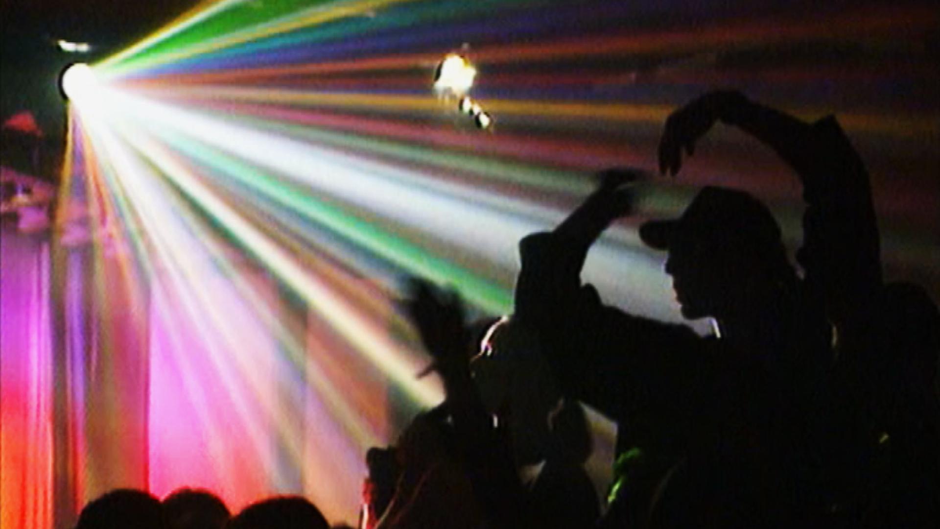 Bunte Lichtstrahlen durchschneiden den dunklen Raum einer Tanzfläche, auf der Silhouetten von Menschen tanzen und sich zur Musik bewegen. Das Bild fängt die dynamische und energetische Stimmung einer lebhaften Party ein.