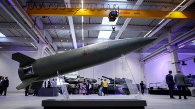 In einer großen Werkshalle ist im Vordergrund eine auf durchsichtigen Stützen aufgestellte Rakete zu sehen. Im Hintergrund stehen Menschen um zwei Panzer.