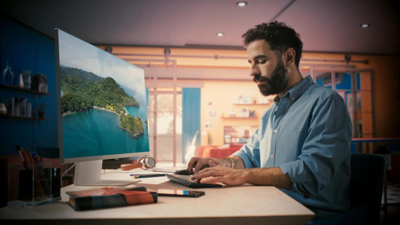 Szene aus "Das große Spiel" von Richard Powers: Seitlicher Blick auf einen Mann, der an einem Schreibtisch mit Bildschirm und Tastatur sitzt. Seine Hände liegen auf der Tastatur. Sein Blick geht auf die Tastur und seine schreibenden Hände. Auf dem Bildschirm ist eine bewaldete Insel mit einer Bucht und einem Sandtstrand zu sehen. Auf dem Schreibtisch befinden sich noch ein Kopfhörer, Kalender und Stifte. Im Hintergrund des Zimmers befindet sich eine Balkontür. An der Wand rechts daneben befinden sich Regalbretter mit Büchern. Der Hindergrund ist weichgezeichnet.