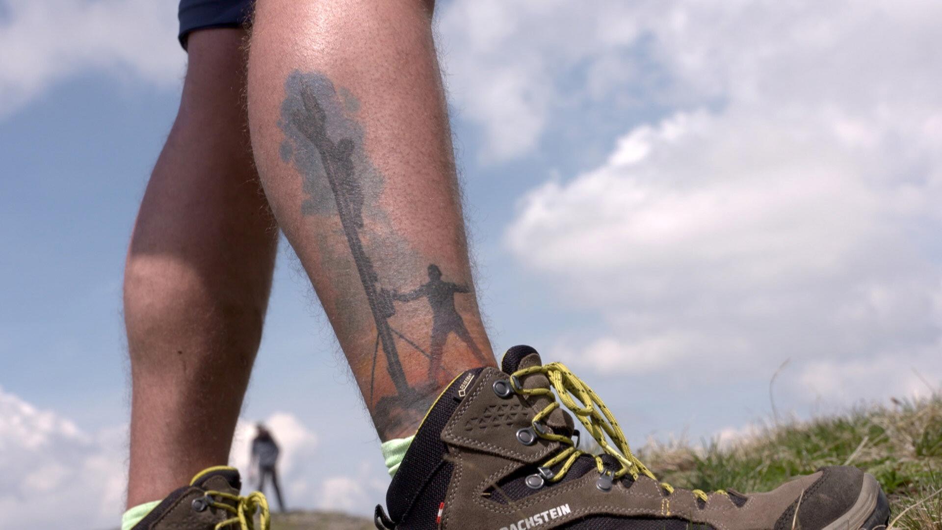Das Bild zeigt das untere Bein eines Menschen, der Outdoor-Schuhe trägt. Die Schuhe sind dunkelbraun mit gelben Akzenten und haben eine robuste Erscheinung, die für Wanderungen geeignet ist. Auf dem untern Teil des Beins ist ein detailliertes Tattoo sichtbar, das eine Silhouette von einer Person zeigt, die an einem Berg oder Baum arbeitet. Das Tattoo hat einen Farbverlauf von rot und orange im unteren Bereich zu schwarz im oberen Bereich, was eine lebendige Darstellung schafft. Im Hintergrund sind einige unscharfe, helle Wolken zu sehen, die auf einen klaren Himmel hinweisen.