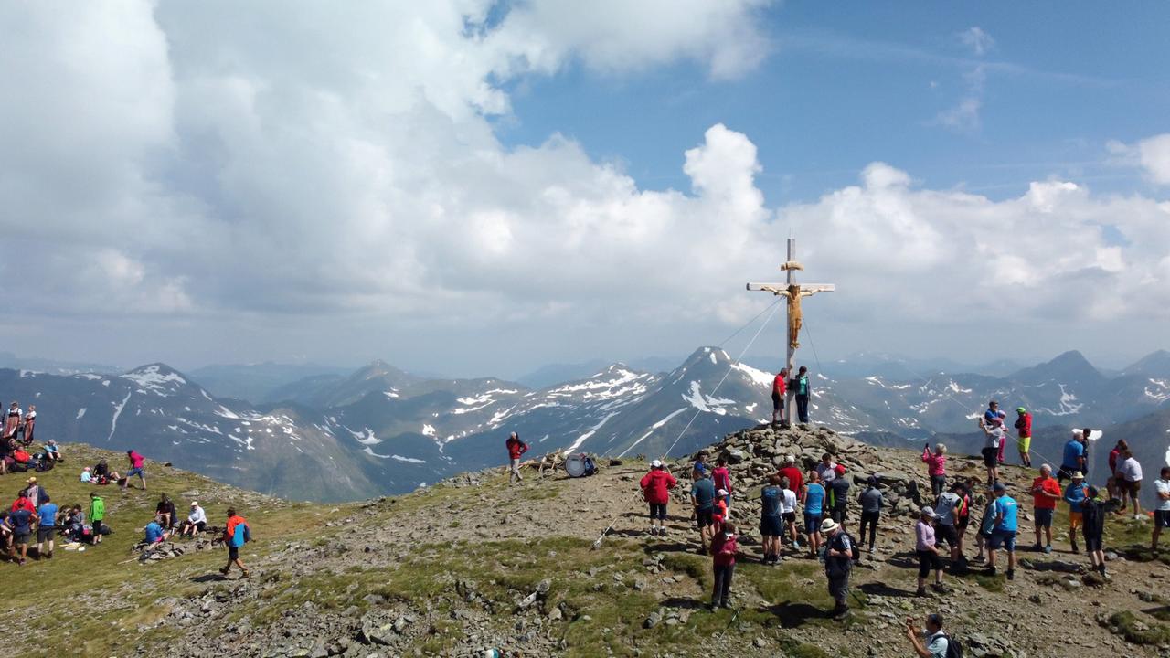 Das Bild zeigt eine Berglandschaft mit einer Gruppe von Wanderern, die sich auf einem Gipfel versammelt haben. Im Vordergrund sind zahlreiche Menschen in unterschiedlichen Farben gekleidet, die teilweise sitzen und sich unterhalten, während andere stehen und die Aussicht genießen. Der Gipfel ist mit grauen Steinen und grünem Gras bedeckt und wird von einem großen, hölzernen Kreuz überragt, das auf einem Steinhaufen steht.  Im Hintergrund erstrecken sich majestätische Berge mit schneebedeckten Spitzen und zerklüfteten Hängen. Der Himmel ist teilweise bewölkt, mit strahlenden Sonnenstrahlen, die durch die Wolken brechen und die Landschaft erhellen. Die Szenerie vermittelt eine Atmosphäre von Gemeinschaft und Naturverbundenheit, während die Wanderer die Schönheit der Umgebung erleben.