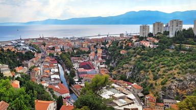 Blick auf eine Stadt, durch die ein Fluss fließt, im Hintergrund Meer und Berge. 