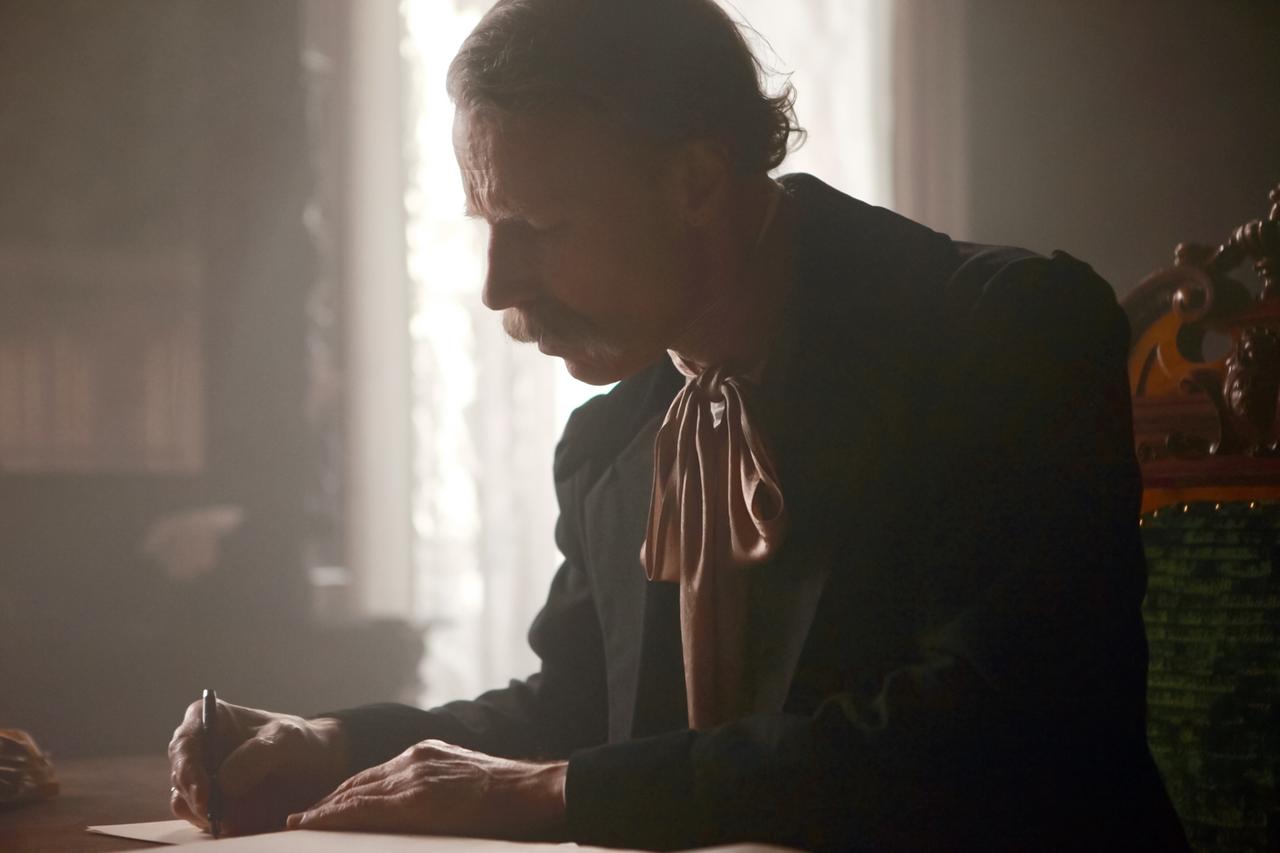 Das Bild zeigt eine historische Szene mit einem Mann, der an einem Tisch sitzt und schreibt. Er hat kurze, lockige Haare und einen Schnurrbart. Er trägt einen schwarzen Anzug mit einem weißen Hemd und einer großen, hellbraunen Schleife um den Hals. Die Beleuchtung im Raum ist gedämpft, mit Licht, das durch ein Fenster auf die Schreibfläche fällt. Hinter ihm sind unscharfe Umrisse von Regalen oder Möbeln zu erkennen. Die Atmosphäre wirkt nostalgisch und erinnert an eine vergangene Zeit.