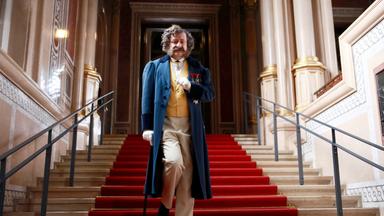 Das Bild zeigt eine männliche Figur, die auf einer beeindruckenden Treppe mit roten Teppichen in einem prächtigen Gebäude herabsteigt. Der Mann trägt einen eleganten blauen Gehrock mit einem gelben Weste darunter. Seine Hose ist hellbeige, und er hat schwarze Schuhe an. Zudem hält er einen Gehstock in der linken Hand und trägt weiße Handschuhe.   Die Umgebung ist reich verziert mit Säulen und prachtvollen Wänden, die möglicherweise aus einem historisch bedeutenden Gebäude stammen. Die Treppe ist aus Stein und führt ins obere Geschoss, wo sich mehr Zierde und vielleicht weitere architektonische Details befinden. Das Licht reflektiert von der Wand, was eine elegante Atmosphäre schafft.   Es scheint sich um eine Szene aus einem historischen Kontext zu handeln, möglicherweise inspiriert durch den Adel oder die aristokratische Gesellschaft, abgebildet im Rahmen einer Dokumentation oder eines Filmes.