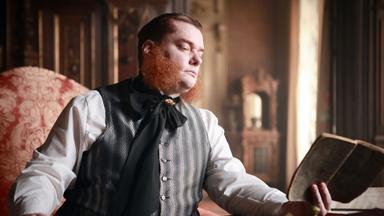 Das Bild zeigt einen männlichen Darsteller mit einem markanten roten Bart und einer informellen, nostalgisch anmutenden Kleidung. Er sitzt an einem Tisch mit mehreren Dokumenten und einer Buchhälfte in der Hand. Die Person blickt nachdenklich zur Seite. Der Hintergrund des Bildes ist eine historisch anmutende Raumgestaltung, die durch elegante Möbel und verzierten Holzarbeiten geprägt ist. Lichteffekte deuten auf einen großen Fensterbereich hin, durch den warmes Licht strömt, was dem Raum eine einladende Atmosphäre verleiht. Der gesamte Stil des Bildes vermittelt eine historische Stimmung.