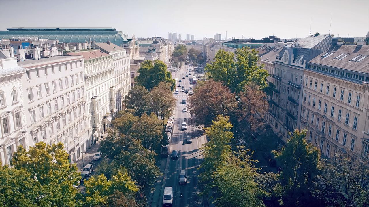 Das Bild zeigt eine Luftaufnahme der Ringstraße in Wien, Österreich. In der Mitte verläuft eine breite Straße, gesäumt von Bäumen, die teilweise herbstliche Farben zeigen. Auf beiden Seiten der Straße sind große, prächtige Gebäude zu sehen, die imarchitektonischen Stil des 19. Jahrhunderts erbaut wurden. Die Fassaden sind teilweise aus hellem Stein, mit großen Fenstern und dekorativen Elementen.  In der Straße sind mehrere Fahrzeuge in Bewegung, darunter Autos und ein Bus. Im Hintergrund erstreckt sich die Skyline von Wien mit modernen Hochhäusern, die sich von den historischen Gebäuden abheben. Das Wetter scheint klar und sonnig zu sein, was eine helle und freundliche Atmosphäre schafft.  Diese Szenerie spiegelt die Geschichte und den Reichtum der Ringstraßenbarone wider, die hier in der Hauptstadt Österreichs einen bedeutenden Einfluss hatten.