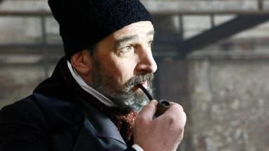 Das Bild zeigt einen Mann in historischer Kleidung, der an einem Tisch sitzt. Der Mann hat einen dichten, grauen Bart und trägt einen schwarzen, filzartigen Hut sowie einen dunklen, eleganten Mantel. Er hält eine Pfeife in der linken Hand und schaut nachdenklich zur Seite. An seinem rechten Hand befindet sich eine Tasse, die auf dem Tisch steht. Der Hintergrund des Bildes ist unscharf und scheint aus alten, gefliesten Wänden oder einer industriellen Umgebung zu bestehen, was auf eine historische Szenerie hindeutet. Die Beleuchtung ist sanft, wodurch das Gesicht des Mannes gut zur Geltung kommt.