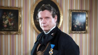 Das Bild zeigt einen Mann in historischer Kleidung, der direkt in die Kamera schaut. Er trägt einen schwarzen Anzug mit einem hellbraunen, gemusterten Weste und einem schwarzen Krawatte. Am Revers seines Anzugs ist eine Auszeichnung mit einer blauen und goldenen Medaille befestigt.   Der Hintergrund besteht aus einer Wand mit vertikalen Streifen in Pink und Cremefarben, an der mehrere Rahmen hängen, in denen Blumen stillleben abgebildet sind. Der Raum hat eine elegante und historische Atmosphäre, die durch die Einrichtung und die Farbwahl vermittelt wird. Es handelt sich um eine Szene, die offensichtlich im 19. Jahrhundert spielt, und setzt einen Fokus auf eine Person von gesellschaftlichem Rang.   Zusammengefasst stellt das Bild eine historische Figur dar, die in einem repräsentativen Raum abgebildet ist, was auf ihre Bedeutung hinweist.