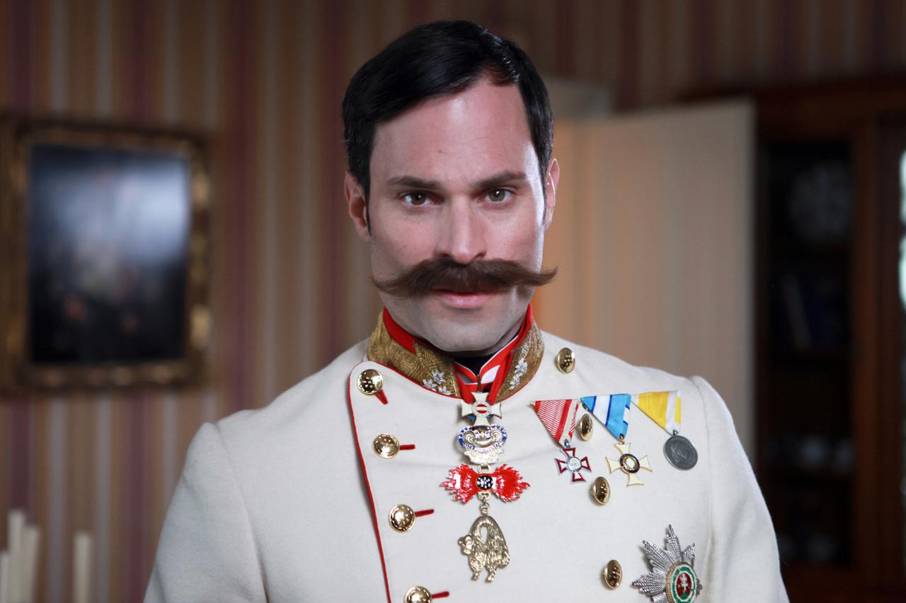 Das Bild zeigt einen Mann in historischer Uniform, die vielschichtige Details und Insignien enthält. Er trägt eine helle Uniformjacke mit goldenen Knöpfen und rot abgesetzten Elementen. An seinem Hals hängt eine Kette mit verschiedenen Orden und Ehrenzeichen, darunter auch ein auffälliges rotes und schwarzes Emblem.   Sein Gesicht ist markant, mit einem kräftigen Schnurrbart und kurzem, glattem Haar. Der Hintergrund ist unscharf, aber deutliche Streifen in sanften Farben sind sichtbar, die eine elegante Innenraumatmosphäre erzeugen. Im Hintergrund sind auch verschwommene objektive Details erkennbar, die auf eine gediegene Räumlichkeit hinweisen.  Das Bild vermittelt einen Eindruck von Autorität und historischer Bedeutung, wahrscheinlich in einem Kontext der Monarchie oder des Militärs.