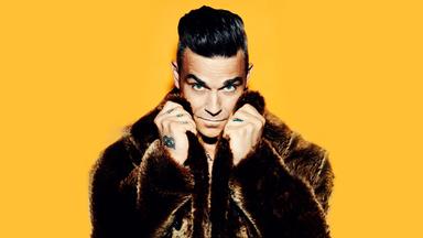 Robbie Williams vor einem gelben Hintergrund. Er trägt einen braunen Fellmantel und hält die Hände am Kragen.