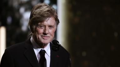 Porträtfoto eines alten Mannes im Smoking, der mit auf dem Rücken verschränkten Armen lächelnd vor einem Mikrofon steht.