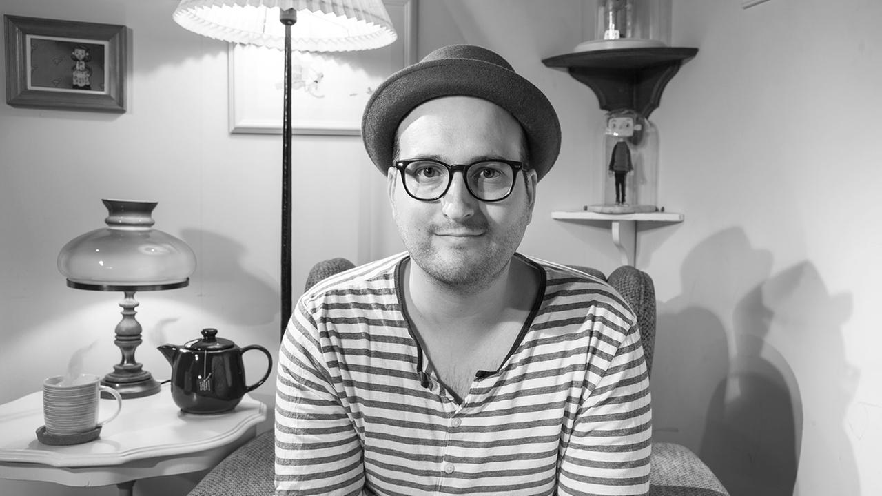 Das schwarz-weiß Portrait eines Mannes mit Hut und Brille in einem Sessel.