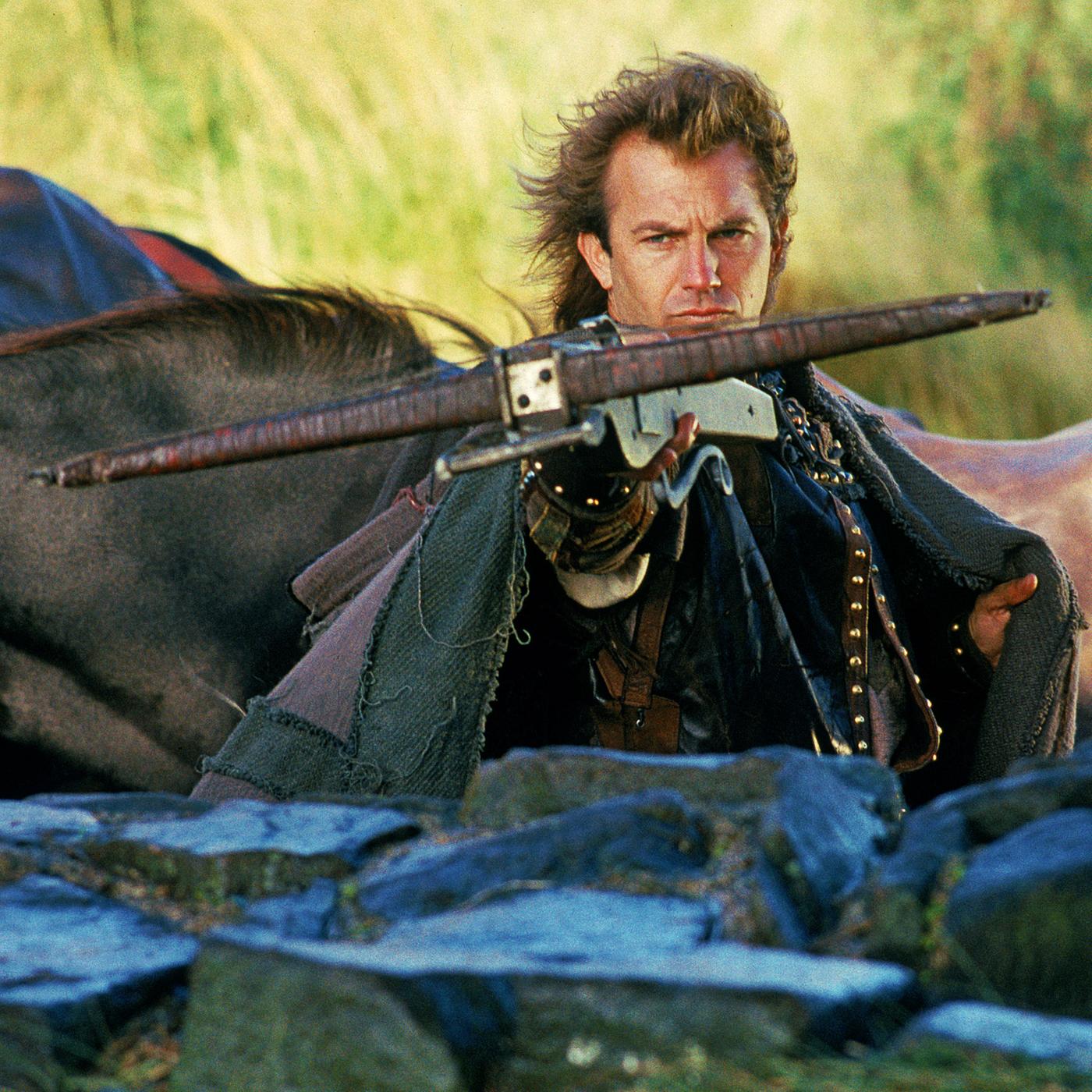 Robin Hood (Kevin Costner) steht hinter einer halbhohen Mauer und zielt mit einer Armbrust nach vorne. Im Hintergrund sind Pferde zu sehen.