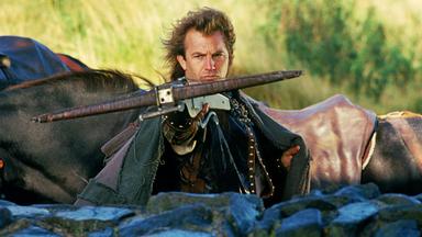Robin Hood (Kevin Costner) steht hinter einer halbhohen Mauer und zielt mit einer Armbrust nach vorne. Im Hintergrund sind Pferde zu sehen.