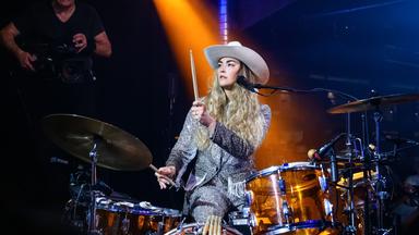 Eine Frau in Pailetten-Anzug und Cowboyhut spielt Schlagzeug.